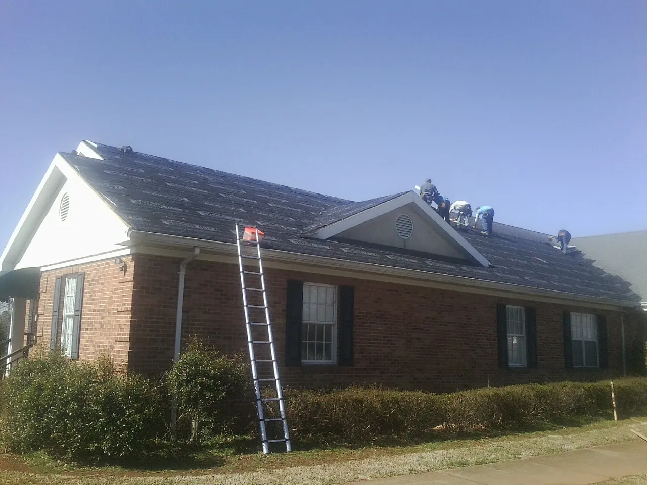 Crew tearing shingles off a brick ranch roof with a ladder set — Metal Roof Repair work by our crew in De Graff
