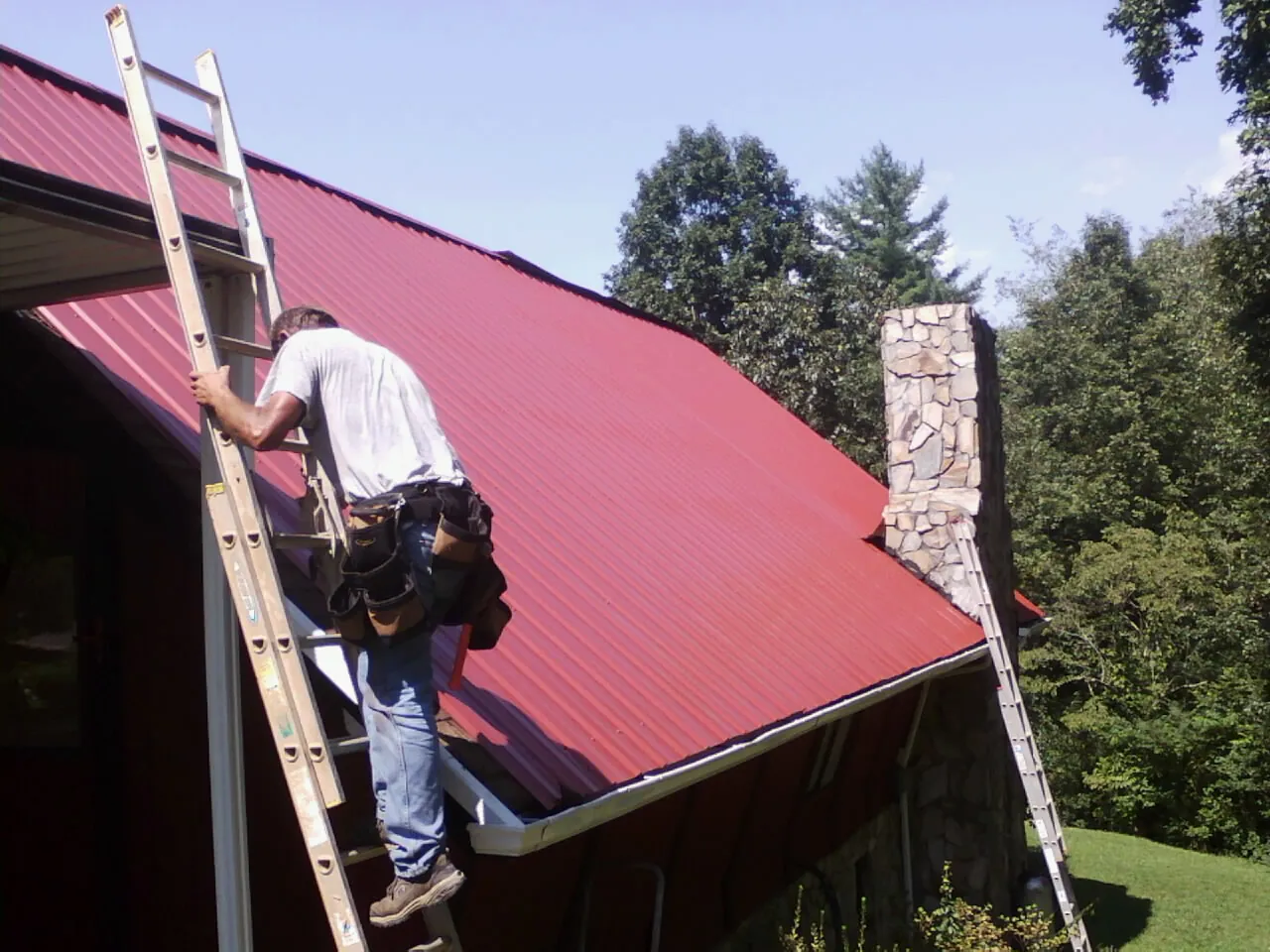 Roofer on a ladder beside a red metal roof and stone chimney — Soffit & Fascia Repair by Roofing Brothers in De Graff